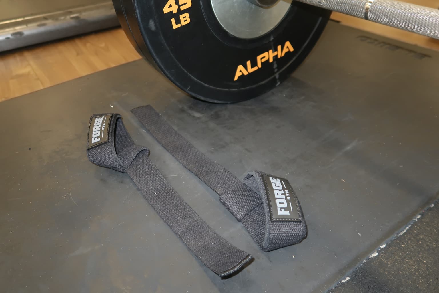 FORGE wrist straps laid out on a gym mat beside a loaded barbell.